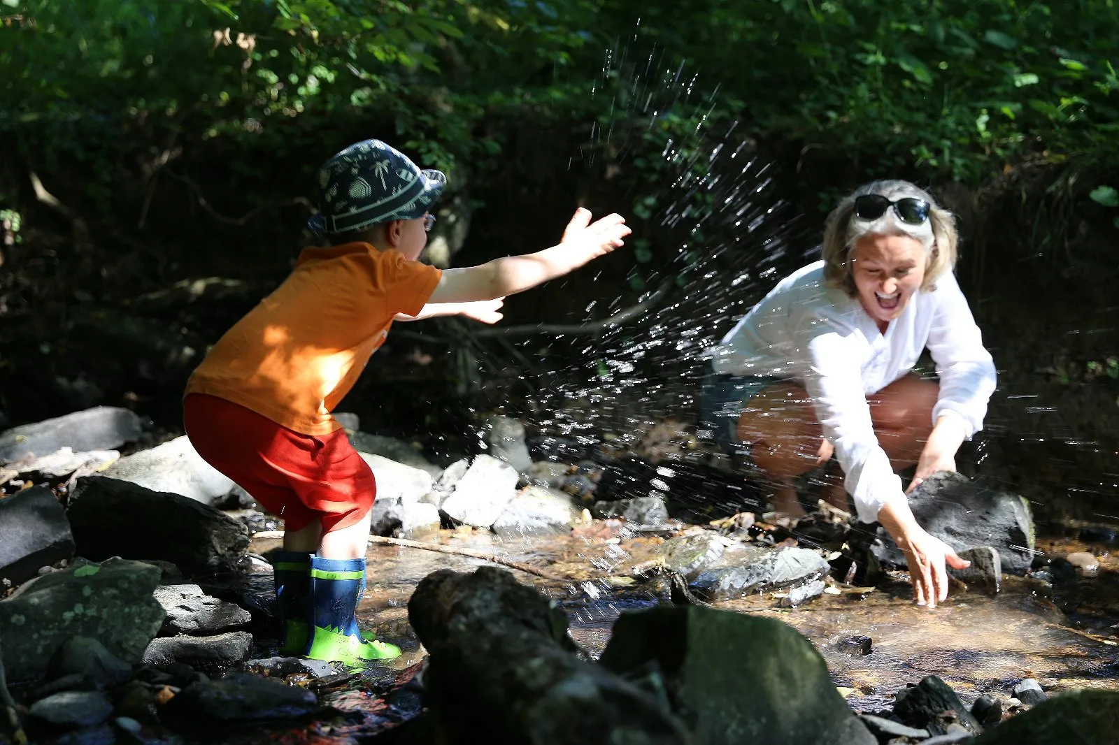 Mi Casa-Dein zu Hause - Familien- und Seniorenbetreuung - Ein kleines Kind in einem orangefarbenen T-Shirt, roten Shorts und einem gemusterten Sonnenhut spritzt Wasser auf eine lächelnde Frau.