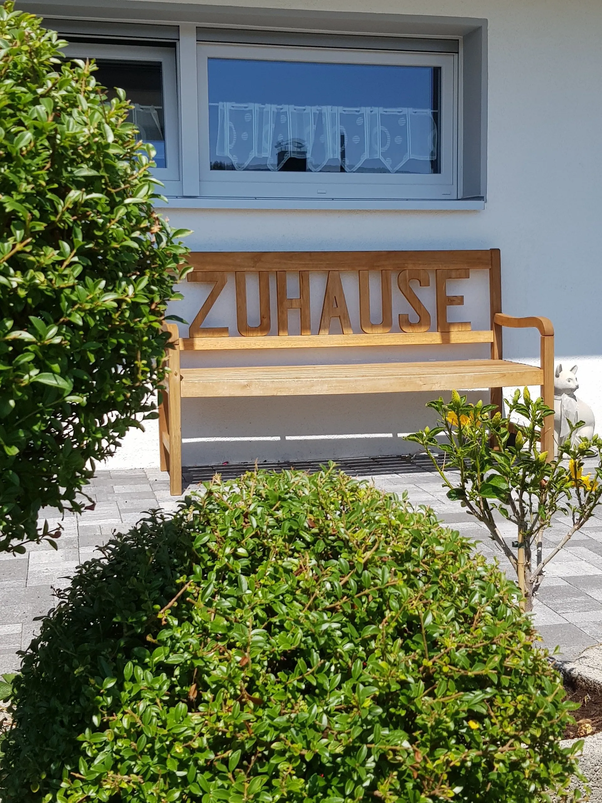 Mi Casa-Dein zu Hause - Familien- und Seniorenbetreuung - Eine hölzerne Gartenbank mit dem in die Rückenlehne eingravierten deutschen Wort „ZUHAUSE“ steht auf einer Steinterrasse vor einem weißen Haus mit großem Fenster. Grüne Büsche rahmen die Szene im Vordergrund und links ein.