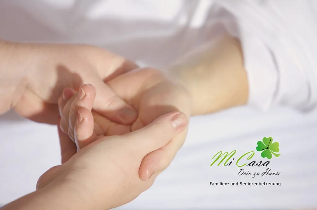 Mi Casa-Dein zu Hause - Familien- und Seniorenbetreuung - Eine Nahaufnahme aus der Vogelperspektive zeigt, wie die Hände einer Person sanft die Handfläche einer anderen Person umfassen und massieren. Beide Personen haben helle Haut, und die Szene ist vor einem weißen, verschwommenen Hintergrund sanft ausgeleuchtet, was ein Gefühl von Ruhe und Geborgenheit vermittelt.