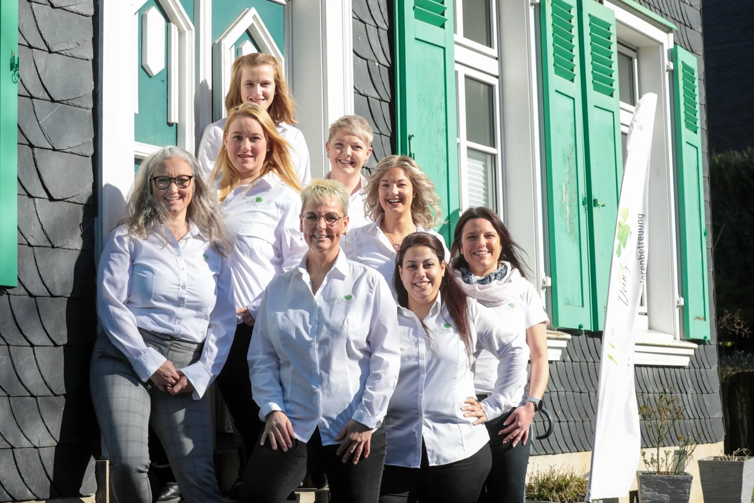 Mi Casa-Dein zu Hause - Familien- und Seniorenbetreuung - Eine große Gruppe von Frauen in weißen T-Shirts posiert für ein Gruppenfoto vor einem Gebäude mit grünen Akzenten.