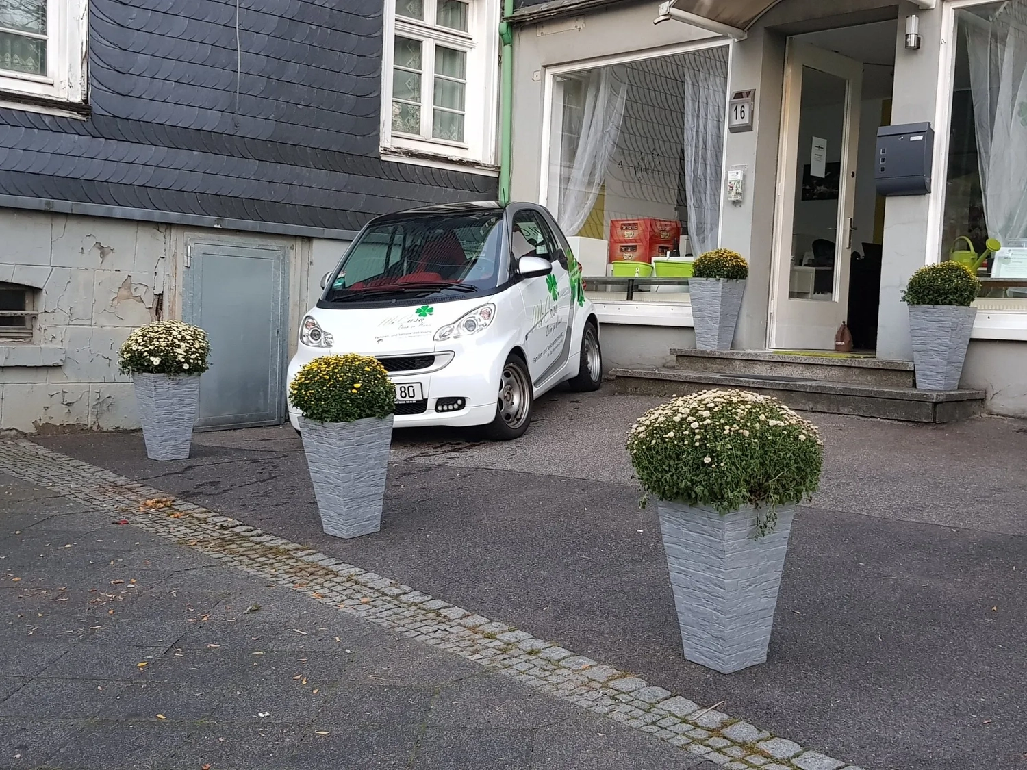 Mi Casa-Dein zu Hause - Familien- und Seniorenbetreuung - Ein kleiner, weißer Zweitürer parkt auf einer gepflasterten Auffahrt vor einem modernen Gebäude mit großen Glasfenstern. Drei hohe, graue Pflanzkübel mit runden, grünen Sträuchern stehen im Vordergrund.