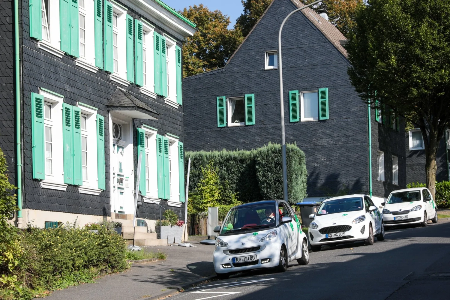 Mi Casa-Dein zu Hause - Familien- und Seniorenbetreuung - Ein weißer Smart parkt in einer schmalen Straße vor traditionellen schiefergedeckten Häusern mit grünen Fensterläden.