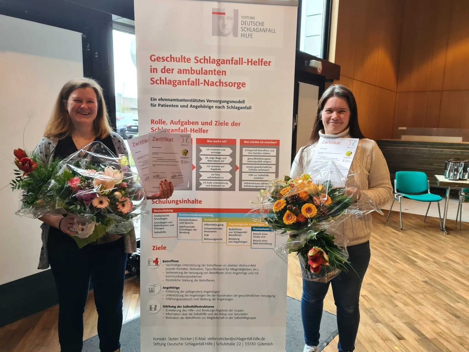 Mi Casa-Dein zu Hause - Familien- und Seniorenbetreuung - Zwei Frauen mit Blumensträußen stehen neben einem Informationsbanner zur Schlaganfallrehabilitation in Deutschland.