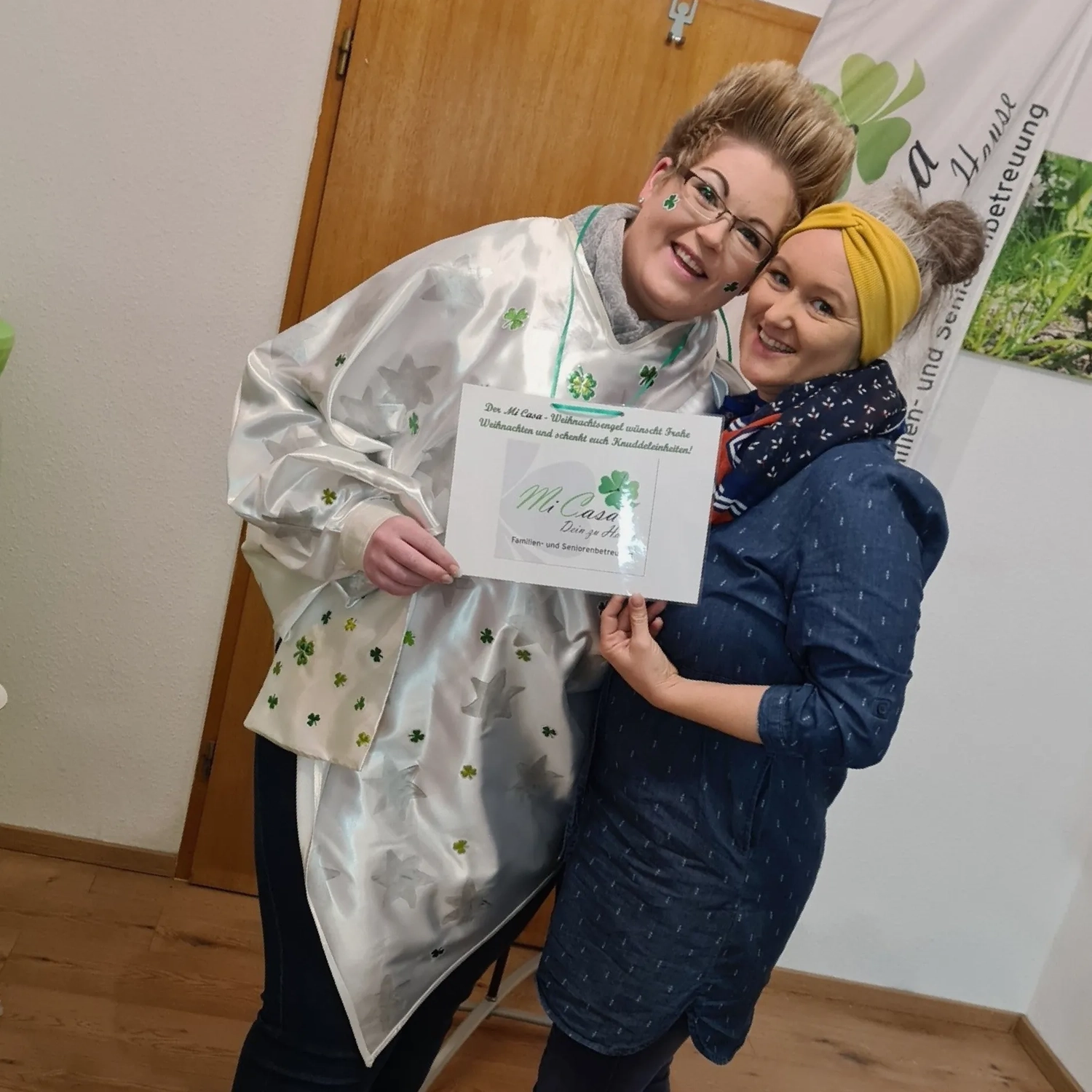 Mi Casa-Dein zu Hause - Familien- und Seniorenbetreuung - Zwei lächelnde Frauen halten eine Urkunde in den Händen; eine trägt einen weißen, gemusterten Umhang, die andere ein blaues Kleid mit gelber Mütze.