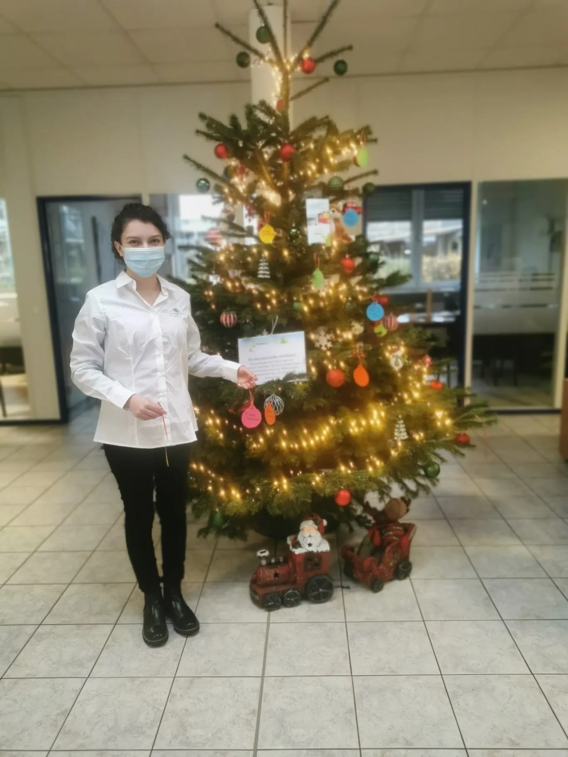 Mi Casa-Dein zu Hause - Familien- und Seniorenbetreuung - Eine Frau in weißem Hemd und mit Mundschutz steht neben einem geschmückten Weihnachtsbaum mit einer Spielzeugeisenbahn am Fuß.