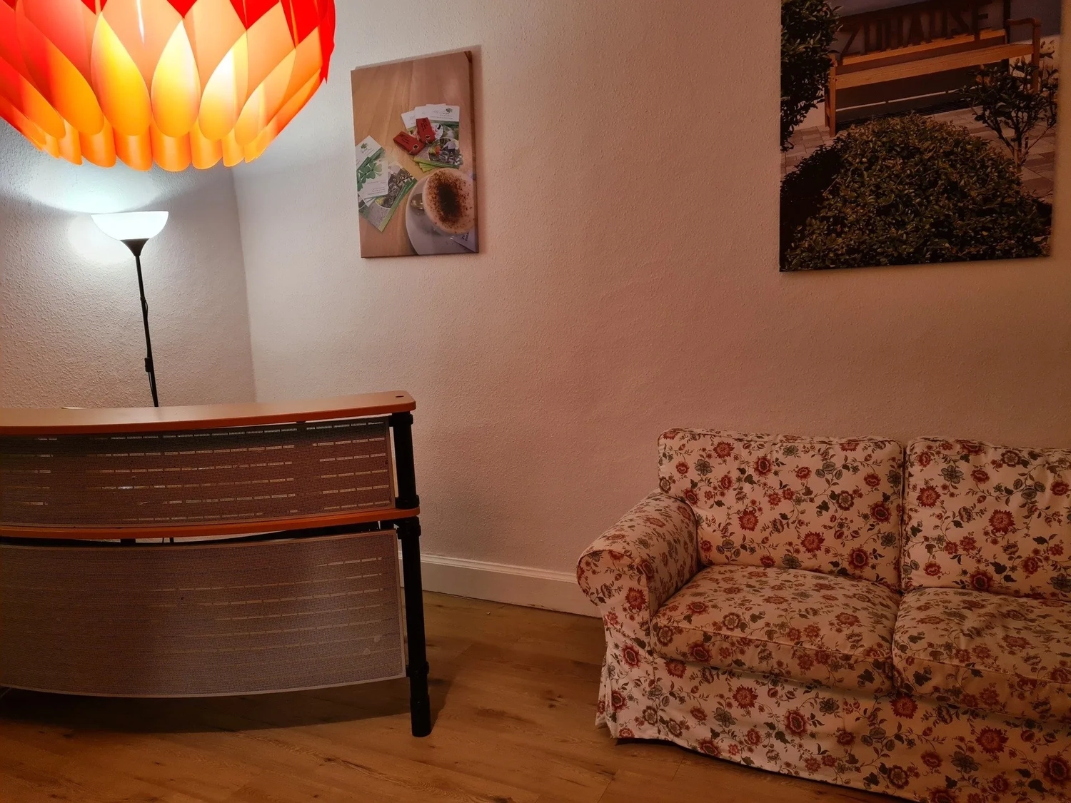 Mi Casa-Dein zu Hause - Familien- und Seniorenbetreuung - Ein gemütlicher Wohnbereich mit einem geblümten Sessel, einem Konsolentisch aus Holz und einer warmen orangefarbenen Pendelleuchte.