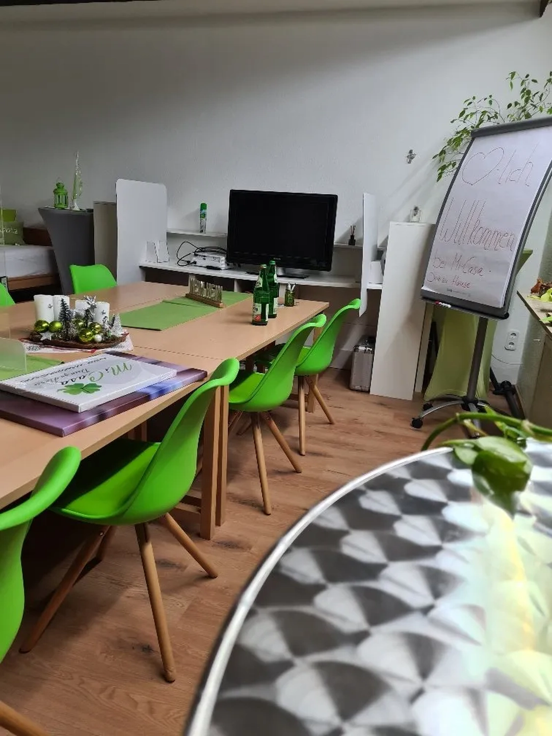 Mi Casa-Dein zu Hause - Familien- und Seniorenbetreuung - Ein Besprechungsraum mit hellgrünen Stühlen, einem langen Holztisch und einem Veranstaltungsplakat.