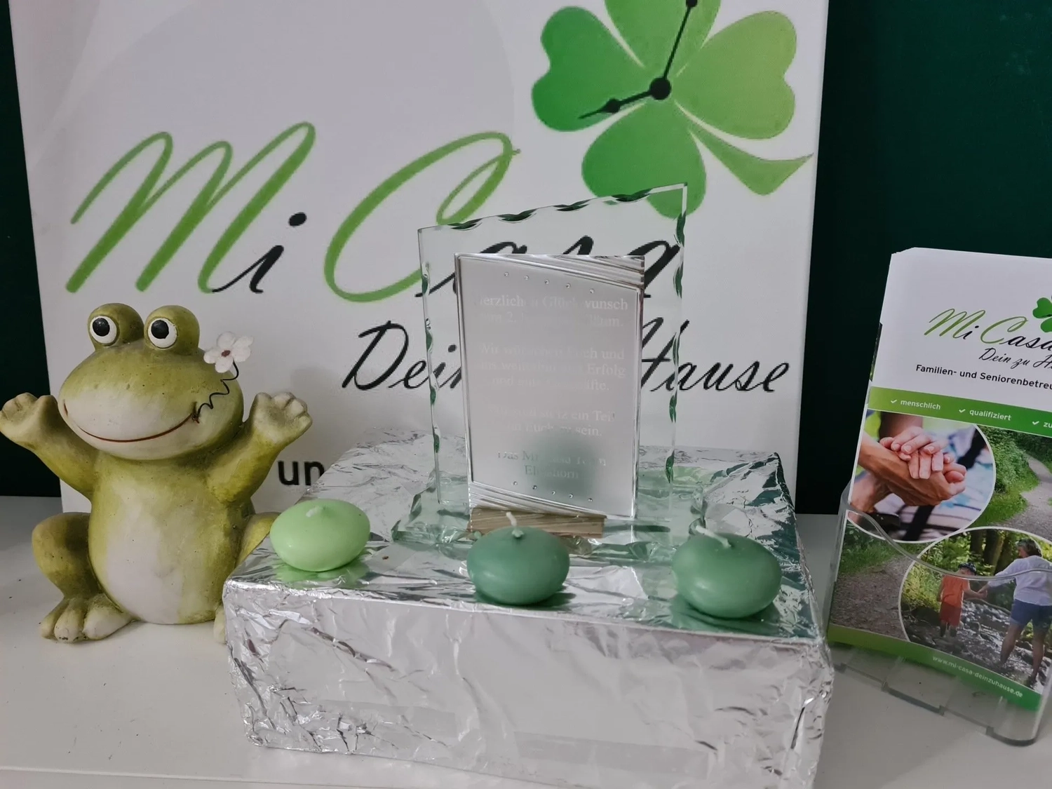 Mi Casa-Dein zu Hause - Familien- und Seniorenbetreuung - Eine Dekoration mit einer Froschfigur, grünen Teelichtern und einem Schild mit einem vierblättrigen Kleeblatt-Logo.