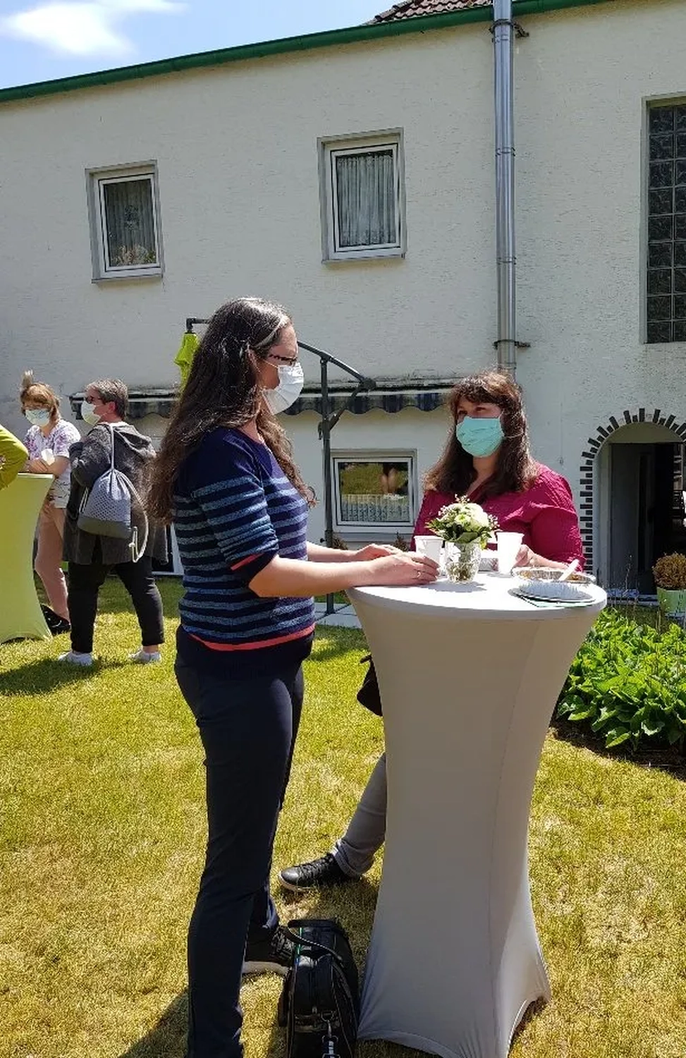 Mi Casa-Dein zu Hause - Familien- und Seniorenbetreuung - Menschen versammeln sich an einem sonnigen Tag im Freien um einen hohen Stehtisch mit weißer Spandex-Abdeckung.
