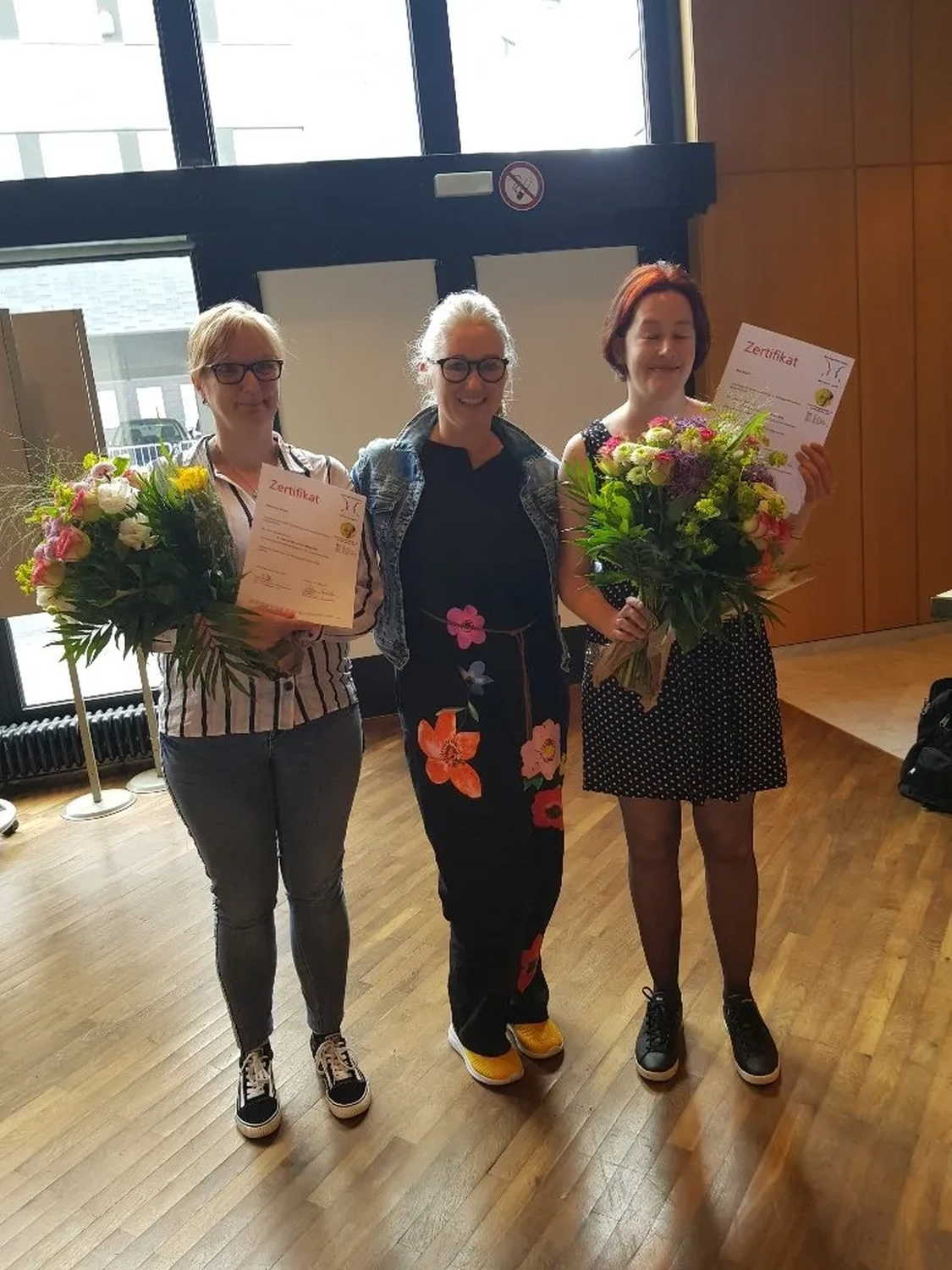 Mi Casa-Dein zu Hause - Familien- und Seniorenbetreuung - Drei Frauen stehen in einem Raum zusammen, zwei von ihnen halten große Blumensträuße und Urkunden.