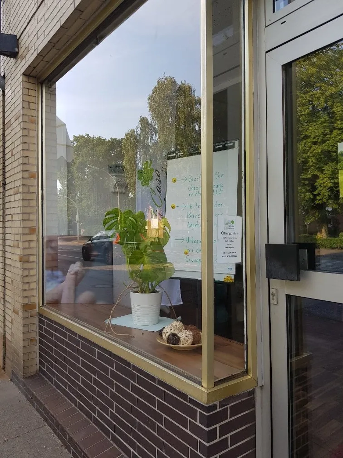 Mi Casa-Dein zu Hause - Familien- und Seniorenbetreuung - Blick auf ein Schaufenster mit Backsteinsockel. Im Schaufenster stehen eine Monstera-Pflanze im Topf und einige Dekorationsgegenstände. Die Spiegelung der Straße ist im Glas zu sehen.