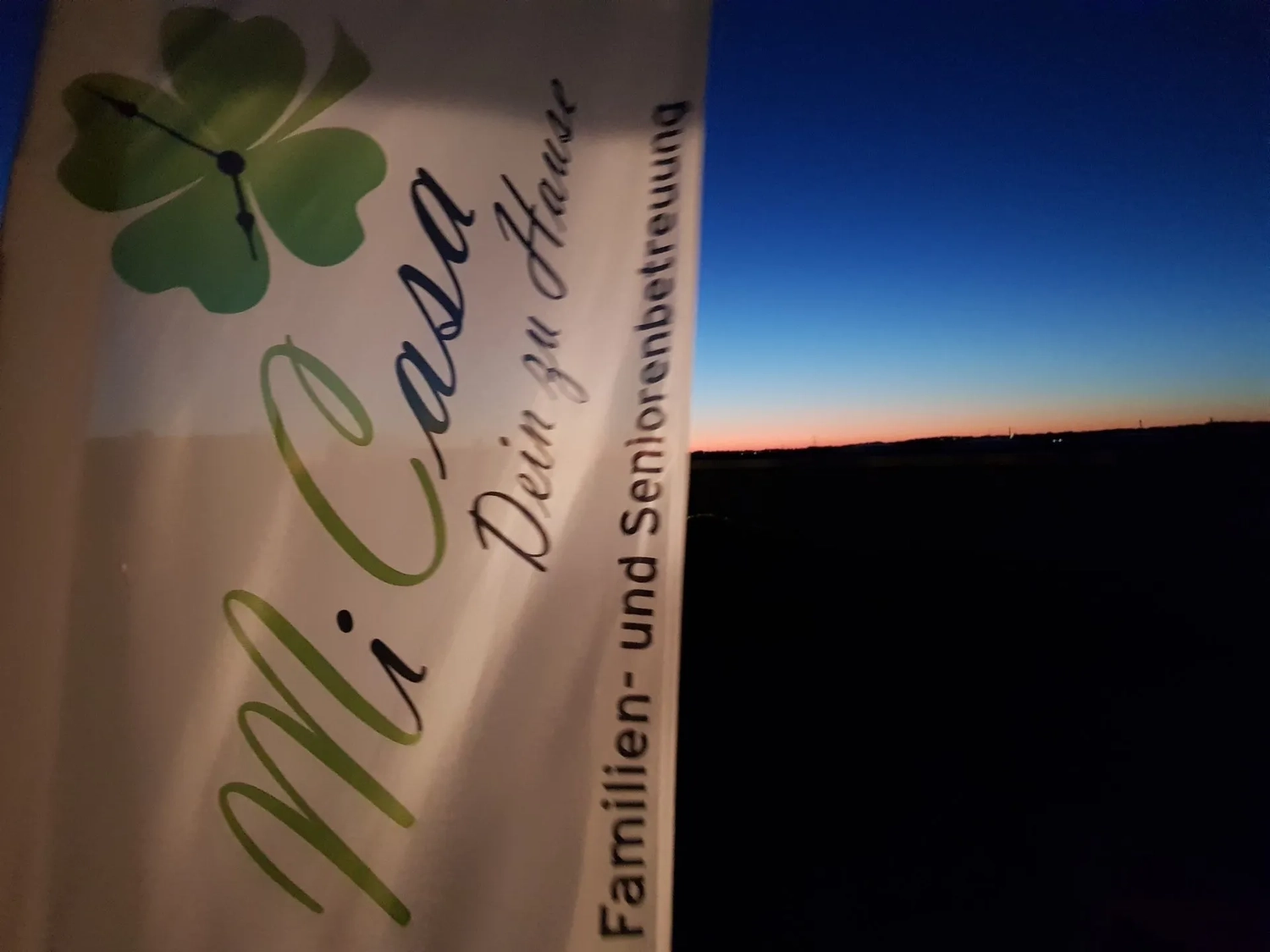 Mi Casa-Dein zu Hause - Familien- und Seniorenbetreuung - Ein vertikales Banner mit der Aufschrift „Mi Casa“ ist vor dem Abendhimmel zu sehen. Auf dem Banner steht unter anderem „Dein zu Hause“ und „Familien- und Seniorenbetreuung“.