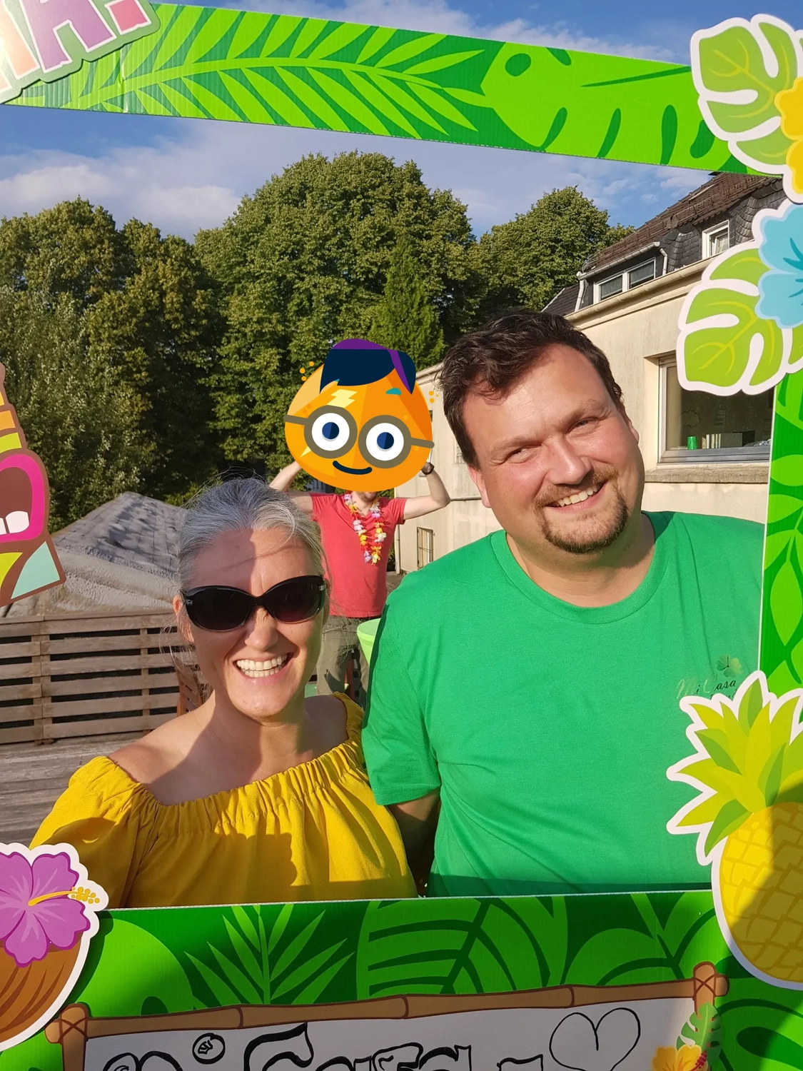 Mi Casa-Dein zu Hause - Familien- und Seniorenbetreuung - Ein Mann und eine Frau posieren hinter einem tropisch dekorierten Fotokabinenrahmen mit Ananas und Blättern. Der Mann trägt ein hellgrünes T-Shirt, die Frau ein gelbes schulterfreies Top und eine Sonnenbrille.