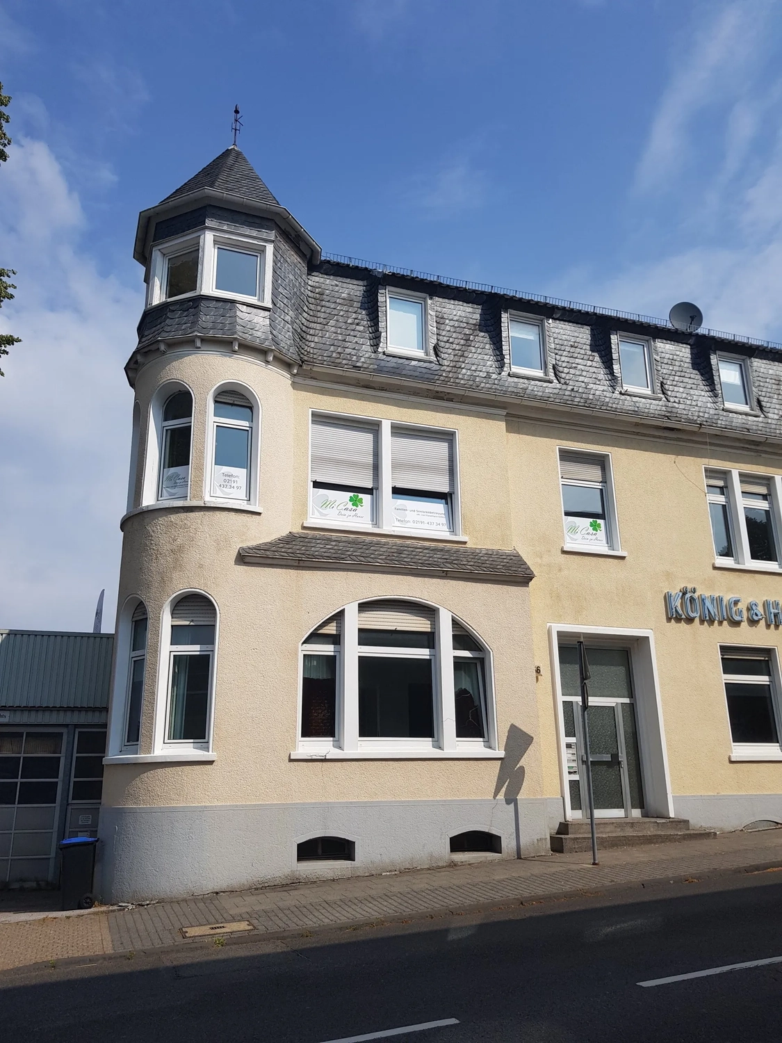 Mi Casa-Dein zu Hause - Familien- und Seniorenbetreuung - Ein zweistöckiges, cremefarbenes Eckgebäude mit einer markanten, geschwungenen Fassade. Es hat große, weiß gerahmte Fenster und eine kleine Eingangstür. Am unteren Bildrand verläuft eine gepflasterte Straße.